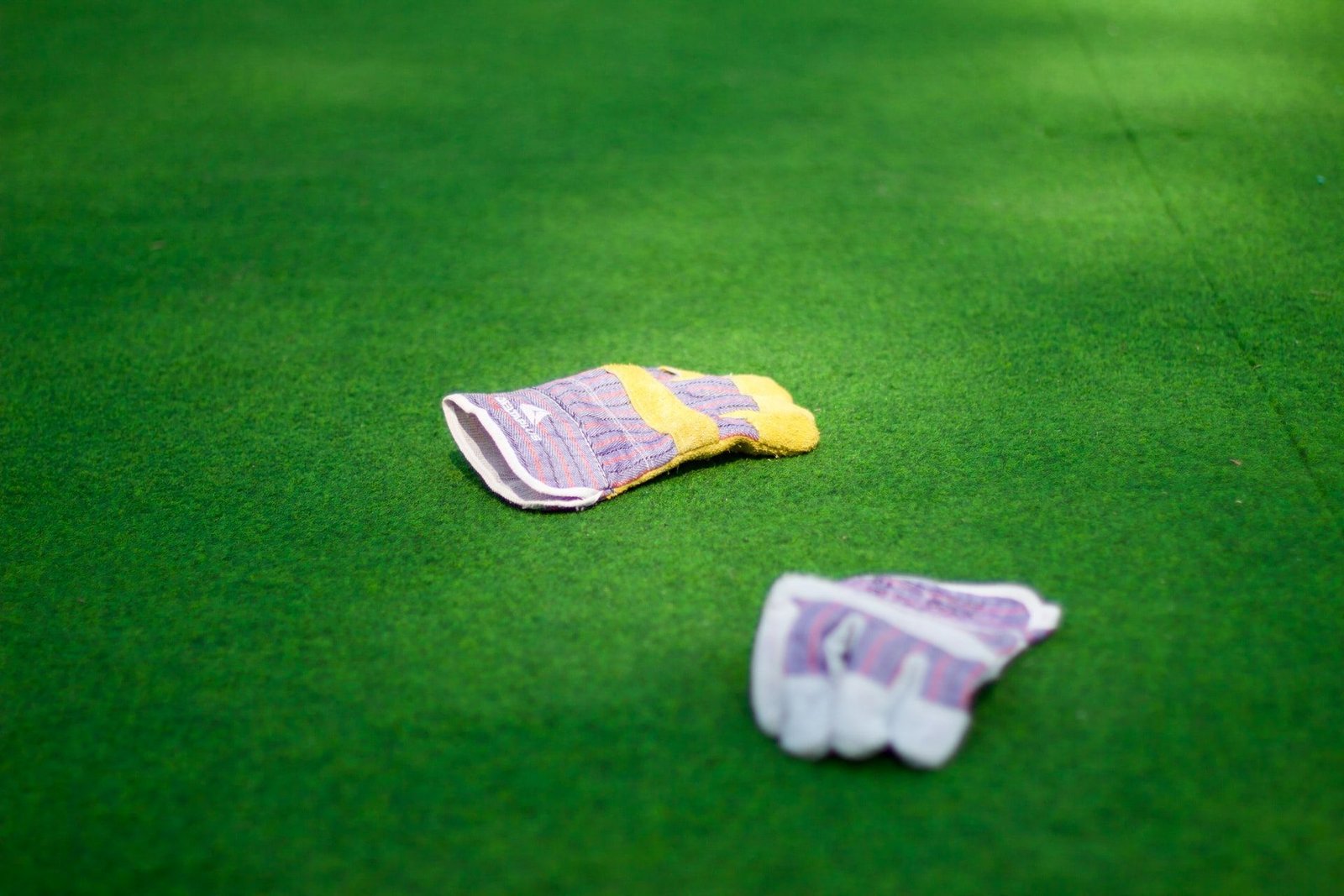 Two work gloves are lying on green grass. One glove is upright with the palm facing down, while the other is slightly crumpled. The gloves are mostly yellow with a purple and white patterned fabric on the cuffs and the backs of the hands, blending seamlessly against the expertly installed turf by Tampa Turf Solutions.