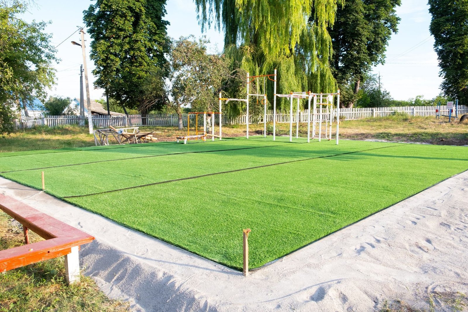 A sports field with artificial grass, expertly laid by dedicated turf installers, surrounded by a sandy border. Situated in a park with trees and a white picket fence in the background, this inviting outdoor space boasts outdoor fitness equipment, picnic tables nearby, and a red bench in the foreground.