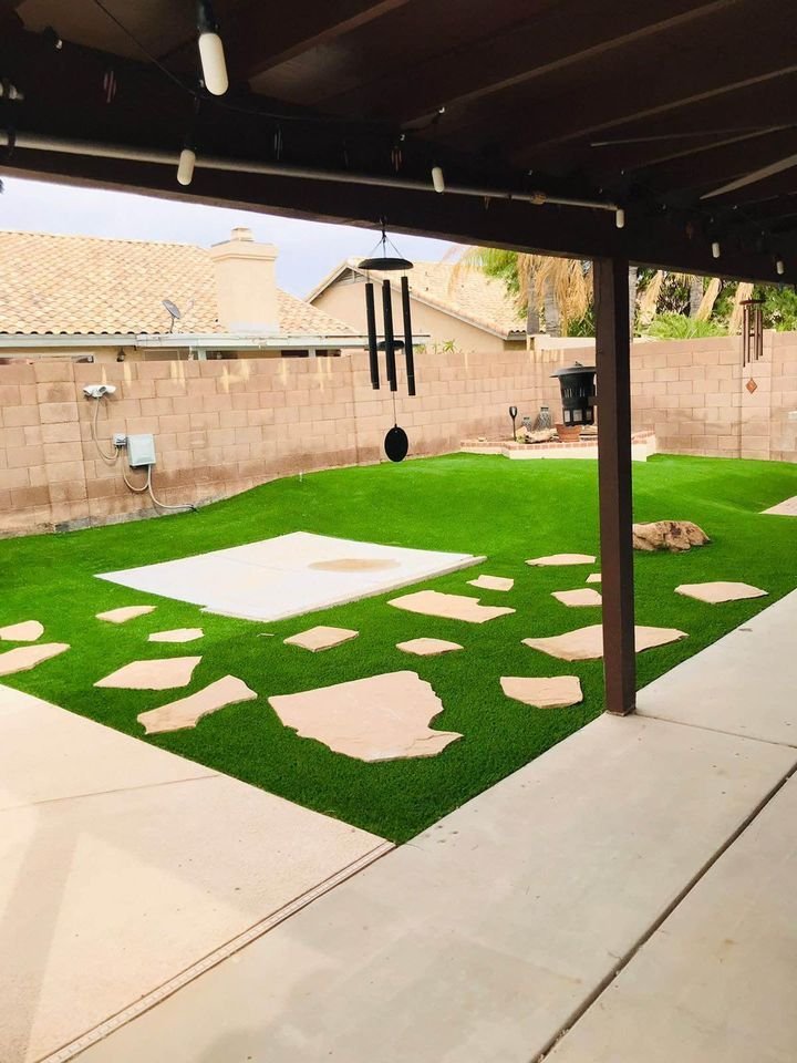 A backyard in Brandon, FL, featuring artificial grass and a concrete slab with scattered large stone stepping stones. A wind chime hangs from the patio roof, and there's a charming stone fireplace. Enclosed by a brick wall, this serene outdoor space also has a dog lounging on the lush turf.