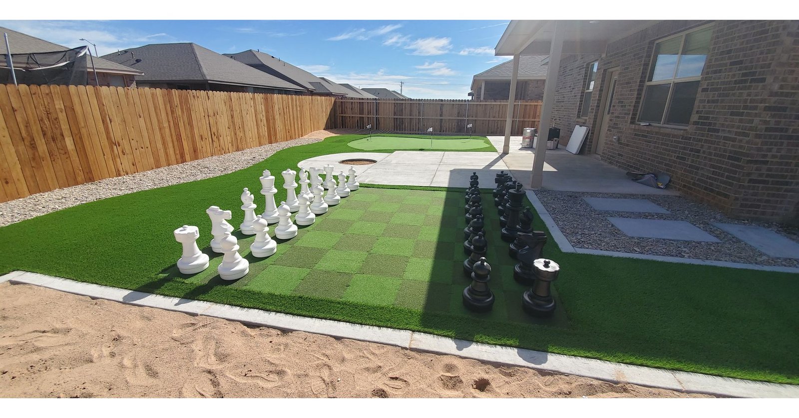 A backyard in Clearwater FL with a life-sized chessboard featuring large black and white chess pieces. Tampa Turf Solutions handled the artificial grass installation, which surrounds the area. The yard also includes a wooden fence, putting green, concrete patio with a covered section, and houses in the background.