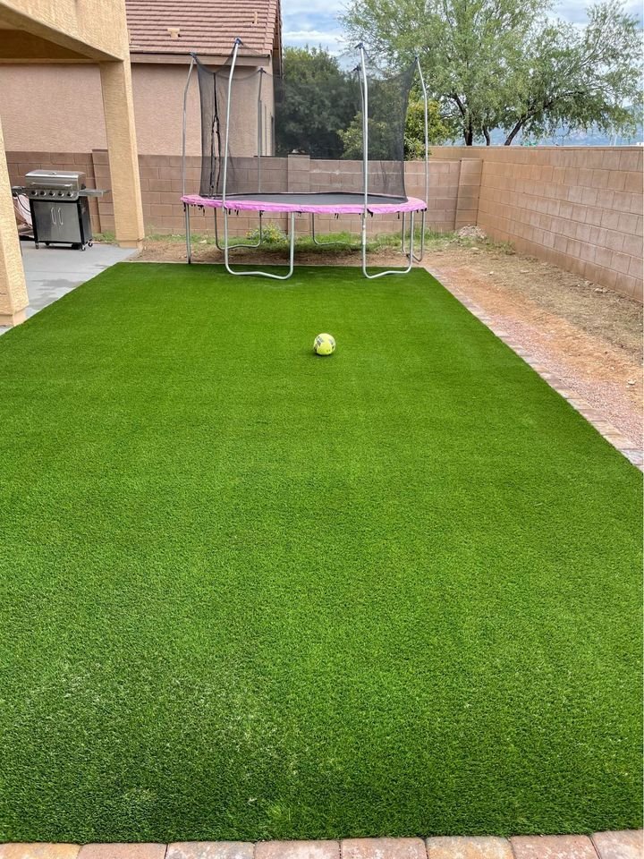 A St. Petersburg outdoor space features a green artificial grass lawn installed by licensed and insured turf pros, a single soccer ball placed in the middle, and a trampoline with a pink border situated next to the house. A barbecue grill is visible on a concrete patio area, all enclosed by a brick wall. Contact us for a free quote!