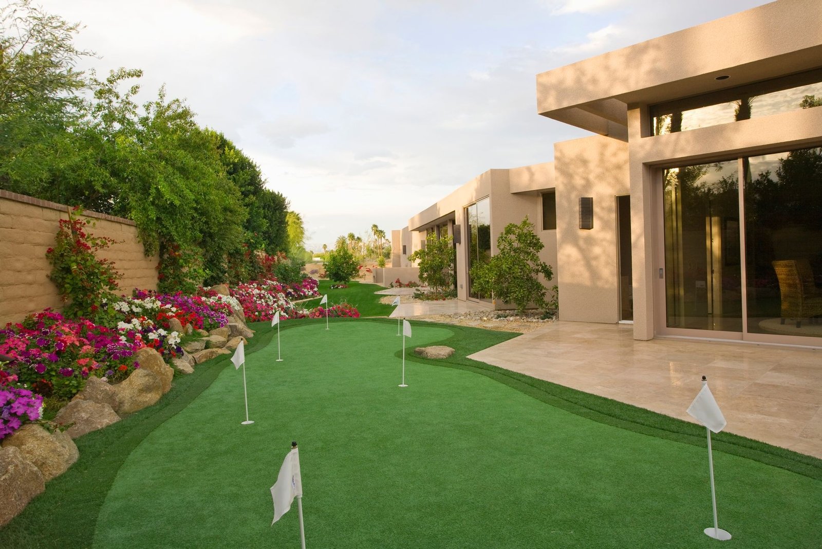 A modern home with large glass doors opens up to a well-manicured St. Petersburg outdoor space featuring a putting green with several holes marked by white flags. Adorned with vibrant flower beds and greenery along a stone pathway, the landscaping was crafted by licensed and insured turf pros.