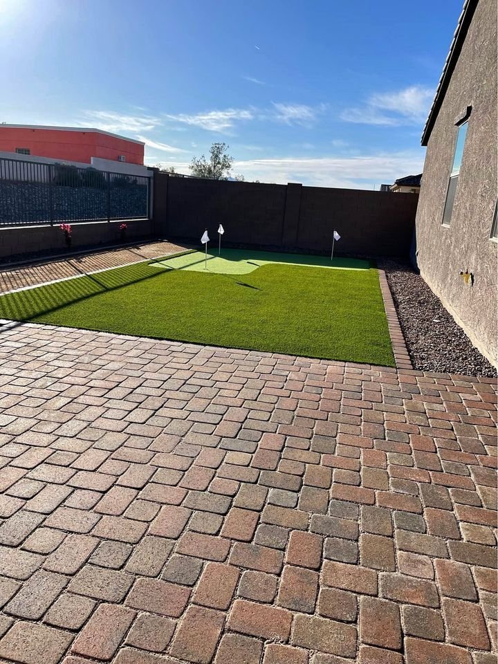 A backyard in Brandon, FL with a brick-paved patio and a small artificial putting green with three holes, each marked by a white flag. The area is surrounded by a low brick wall and has a clear blue sky overhead. Contact us for an artificial turf installation or to get your free quote today!