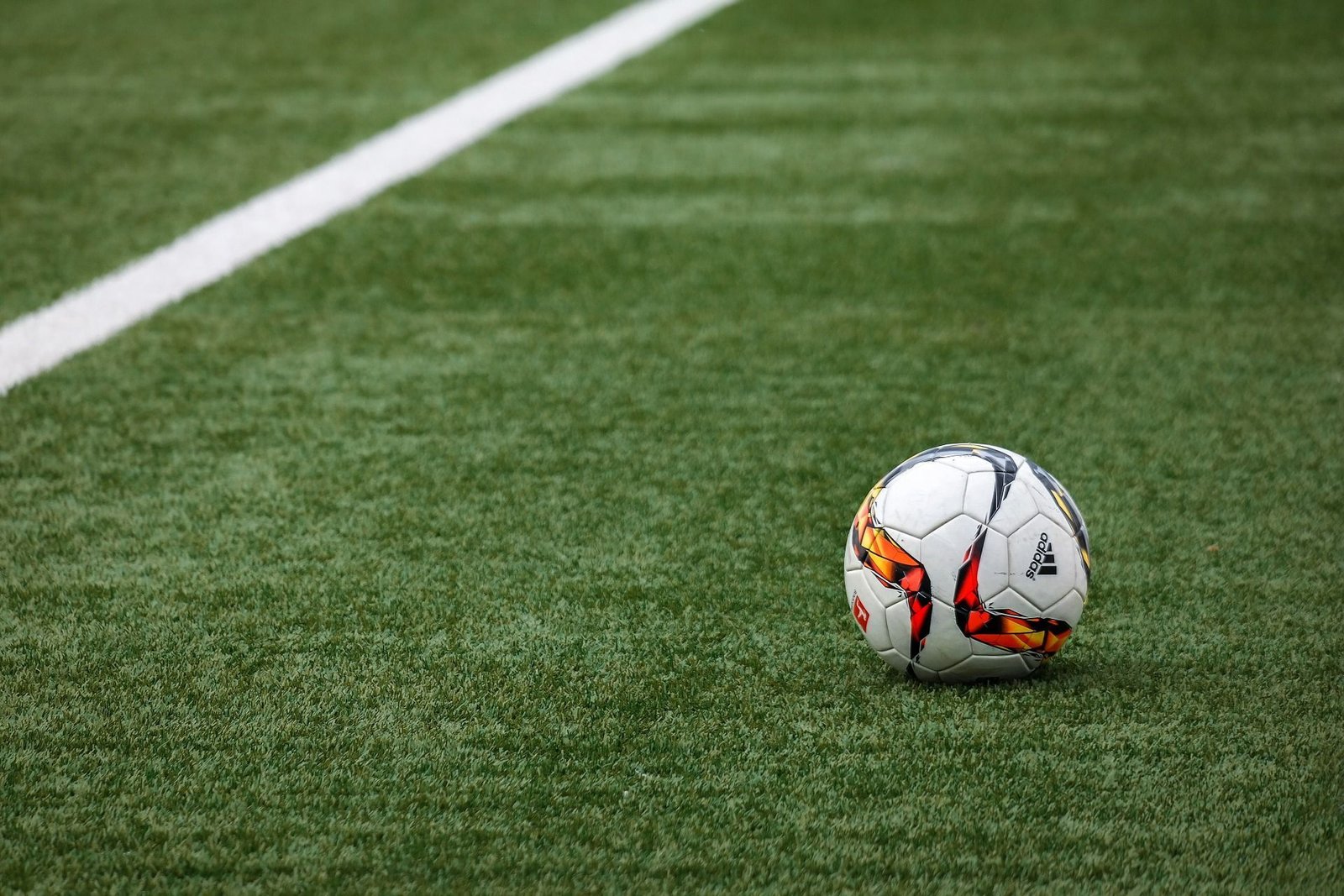 A soccer ball with colorful patterns sits on a green artificial turf field near a white sideline, ready for play. The image focuses on the ball and the clear, bright pet-friendly turf surface.