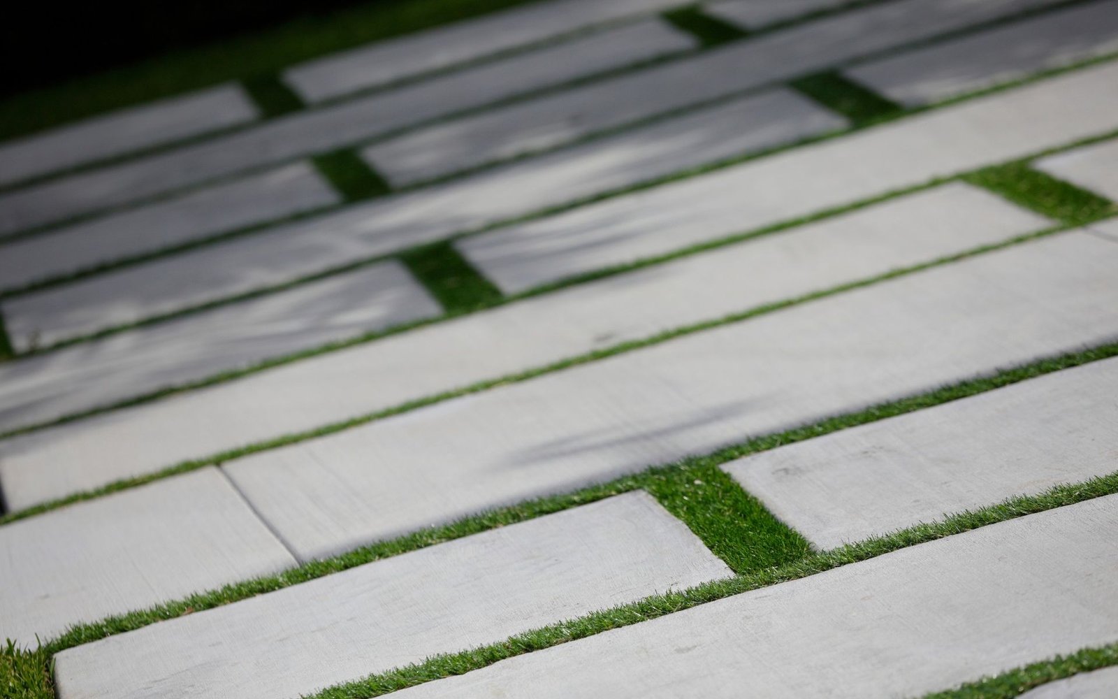 A close-up photo of a pathway composed of rectangular white stepping stones placed in a grid pattern. Green grass, courtesy of professional turf installers, fills the gaps between the stones, creating a visually appealing contrast. Shadows of nearby objects are cast on the pathway in this St. Bradenton outdoor space.