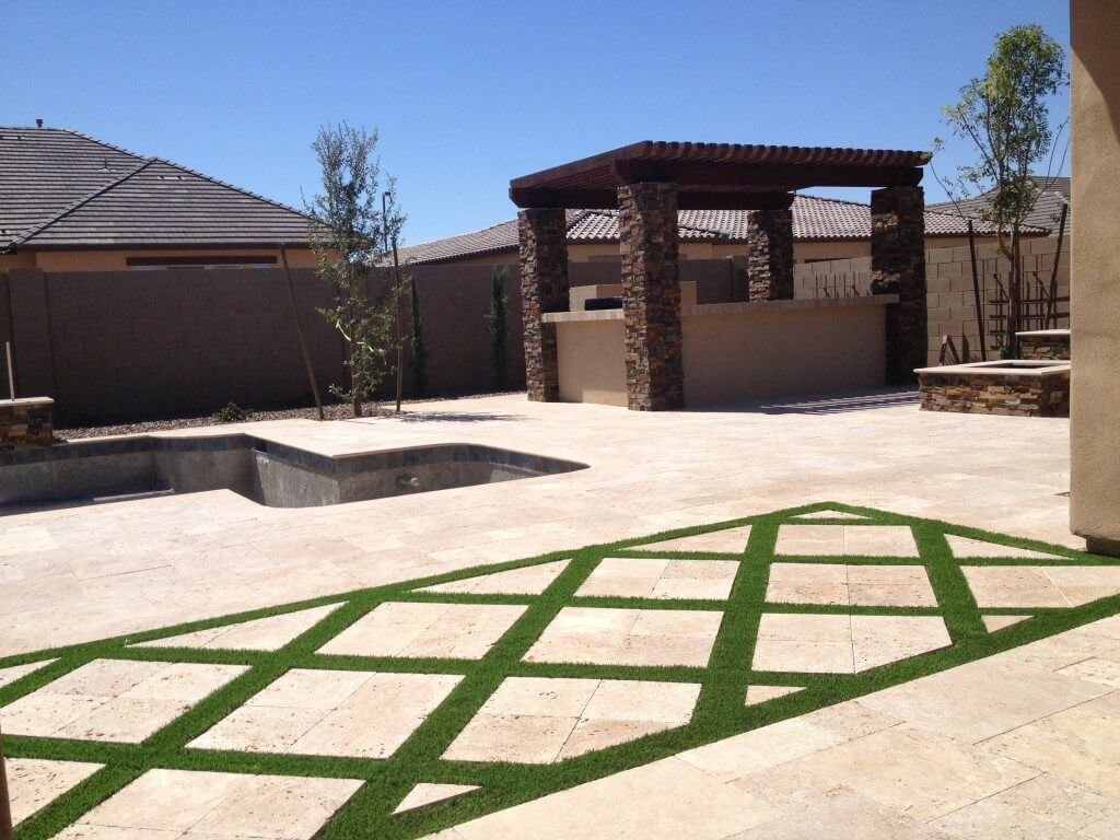 A spacious backyard features a tiled patio area with a diamond pattern of artificial grass inlays. There's a stone-bordered, empty rectangular pool area, and a covered outdoor kitchen with a pergola overhang and stone pillars in the background. Expertly installed by Tampa Turf Solutions, Clearwater FL.
