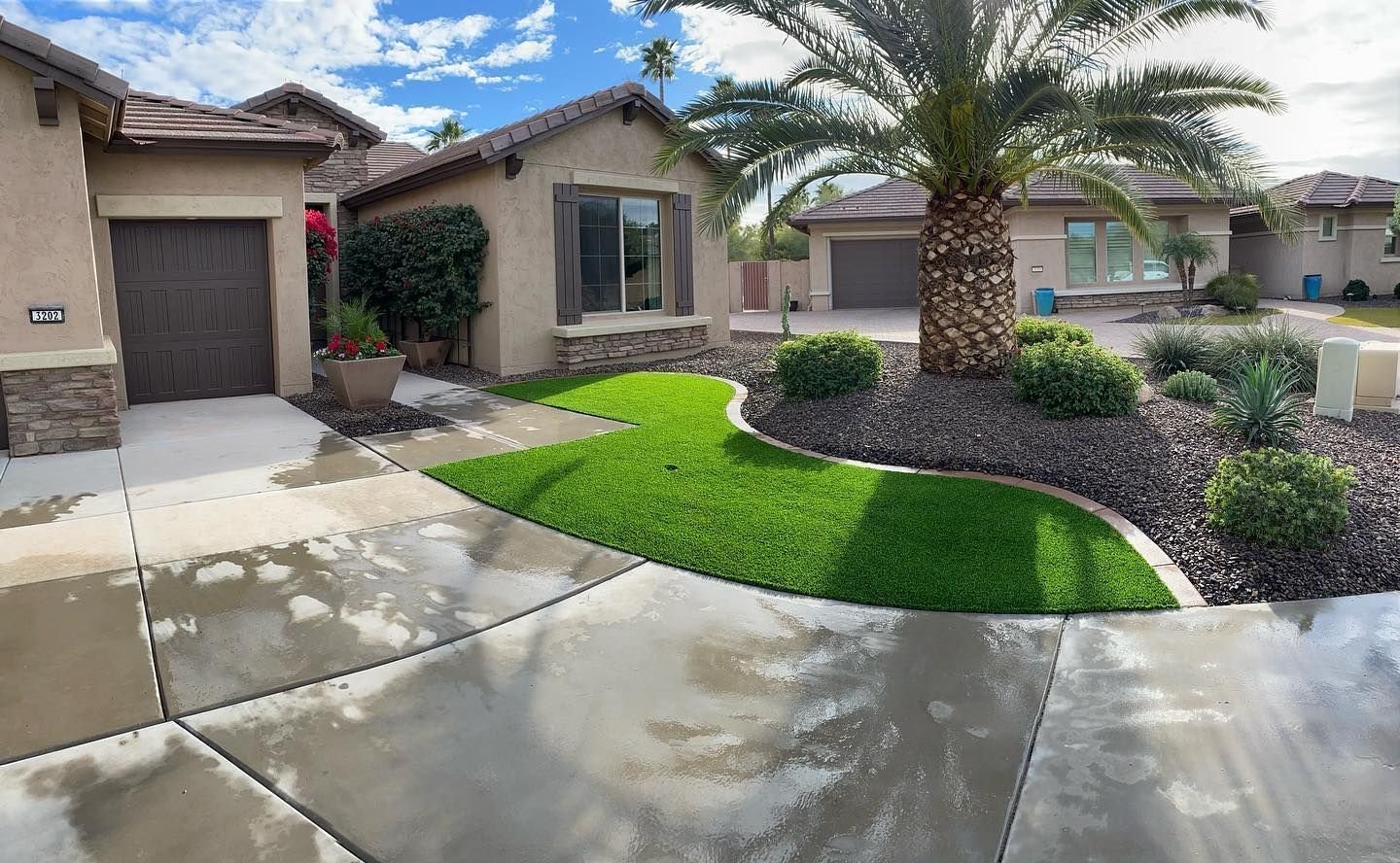 A modern suburban house with beige walls and a brown roof, featuring a well-maintained front yard with green artificial grass installed by licensed turf pros, a palm tree, and desert landscaping. The driveway and walkway are wet, suggesting recent rain or watering. Other similar houses are in the background.